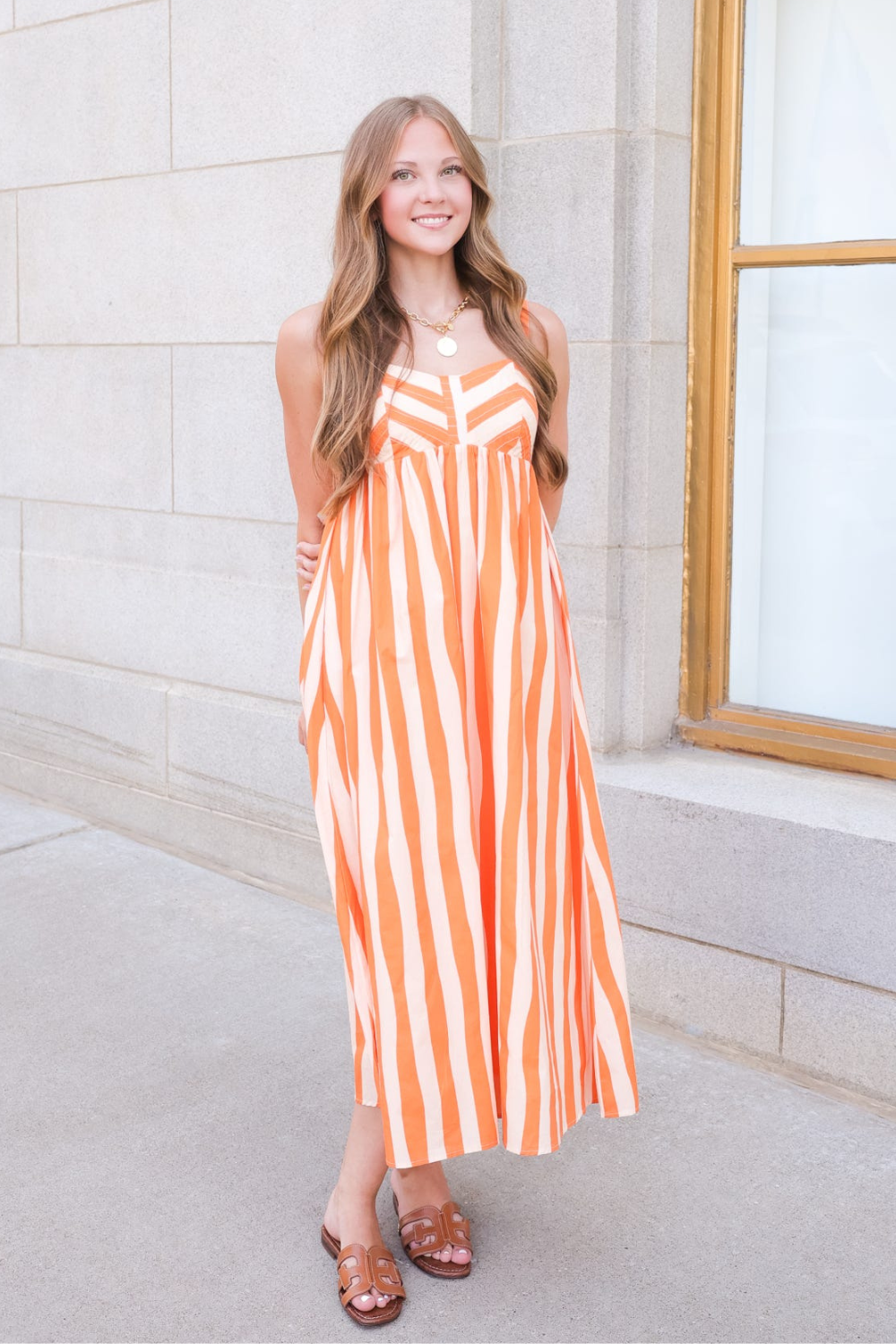 A woman wearing a sleeveless orange and white vertically striped maxi dress and brown strappy sandals, standing outdoors against a light stone building wall.