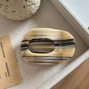 A tortoiseshell hair claw clip with a cream, brown, and black striped pattern, displayed on a textured beige fabric surface next to a brown card.