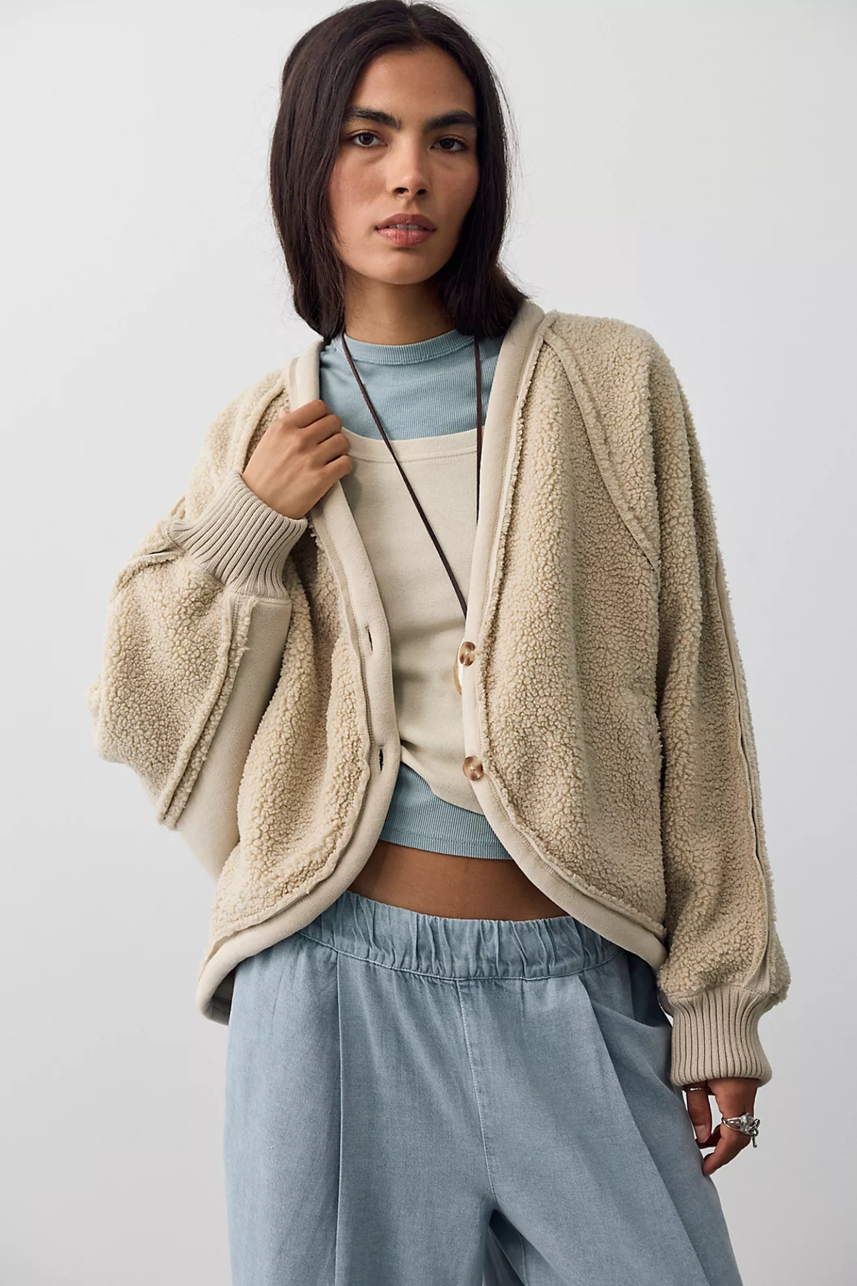 A woman modeling a beige sherpa fleece button-up cardigan over a light blue top and light blue wide-leg pants, photographed against a plain light gray studio background.