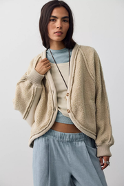 A woman modeling a beige sherpa fleece button-up cardigan over a light blue top and light blue wide-leg pants, photographed against a plain light gray studio background.