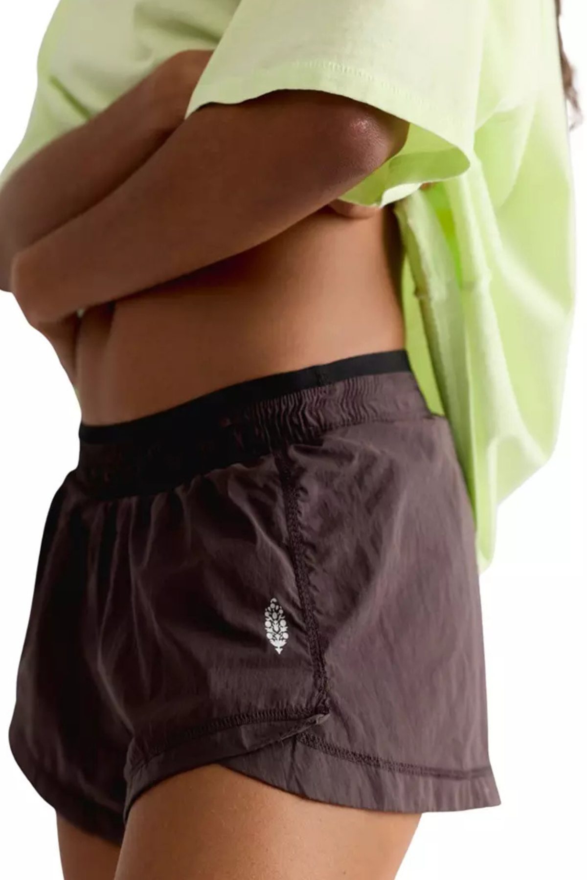 Close-up of a model wearing dark brown athletic shorts with a black elastic waistband and a small silver logo, paired with a neon yellow-green t-shirt against a white background.