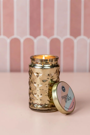 A lit gold-toned glass jar candle with a matching lid leaning beside it on a pink surface, set against a pink striped backdrop.