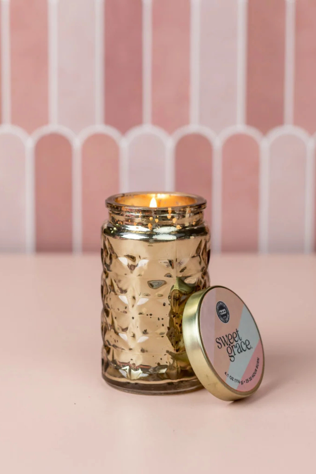A lit gold-toned glass jar candle with a matching lid leaning beside it on a pink surface, set against a pink striped backdrop.
