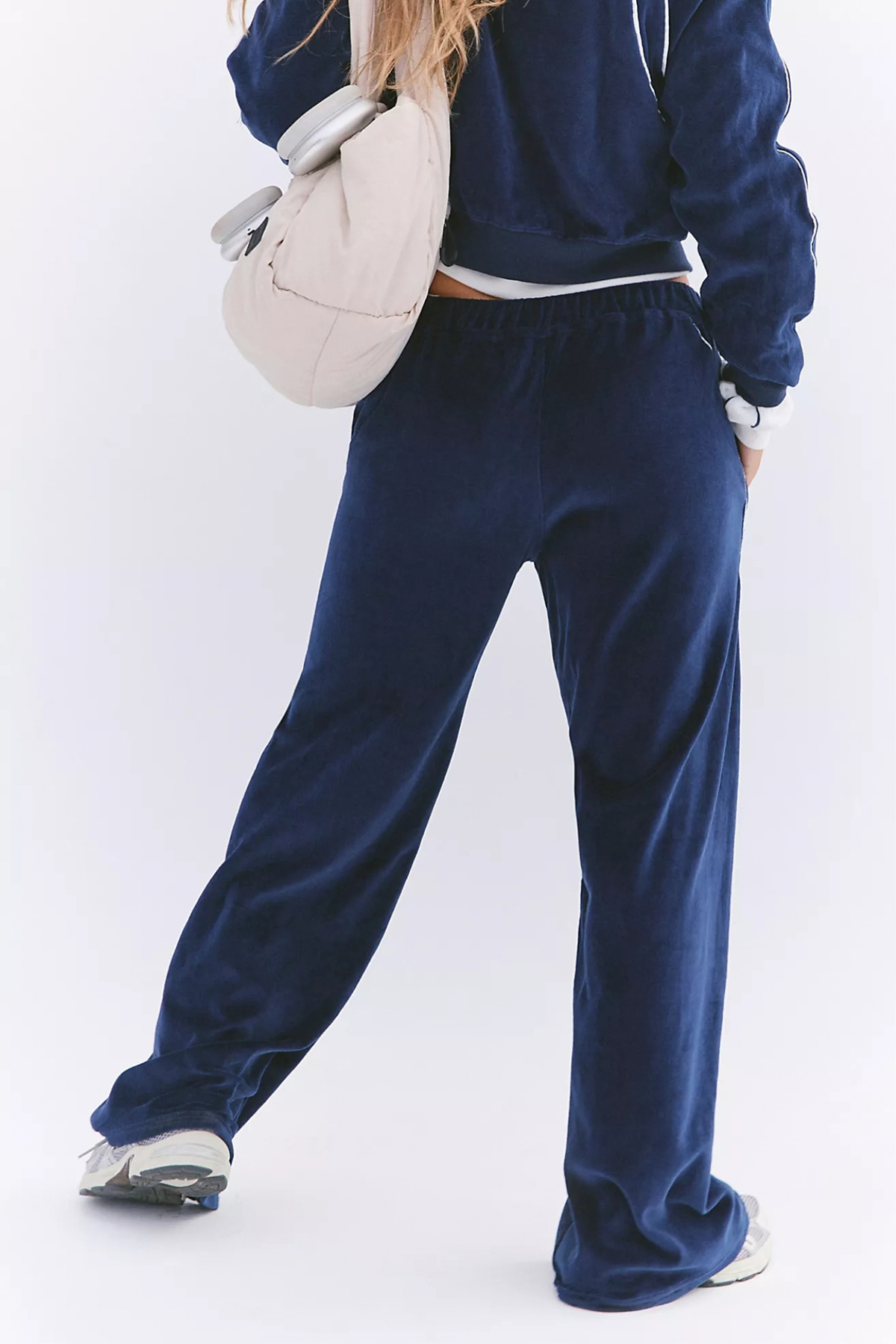 A person wearing a matching navy blue velour lounge set, featuring wide-leg sweatpants and a cropped sweatshirt, styled with a cream shoulder bag and white sneakers against a white studio background.