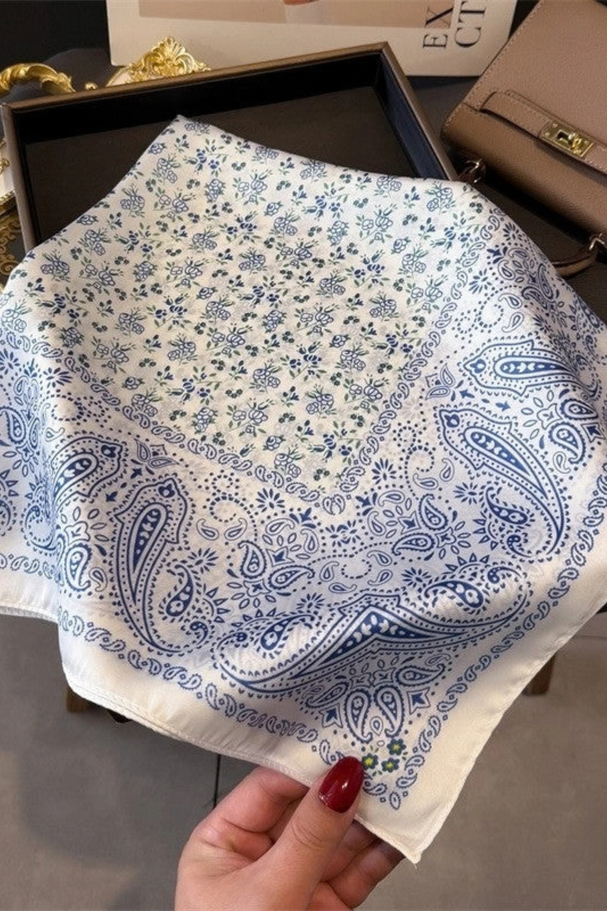 A hand with red nail polish holds a white square bandana featuring a blue paisley and floral pattern, with a dark box and a beige handbag in the background.