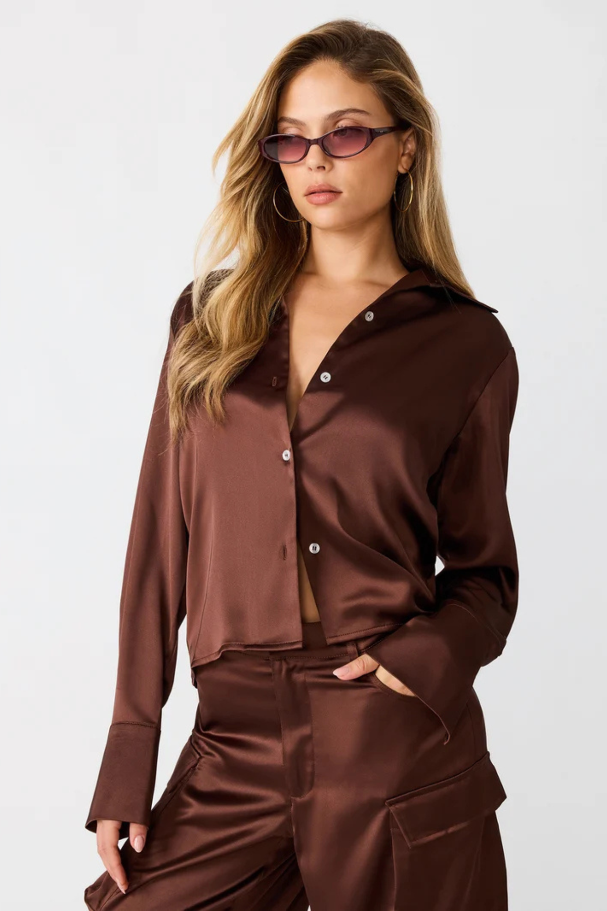 A woman modeling a matching brown satin button-up shirt and high-waisted cargo pants set against a plain light gray studio background.