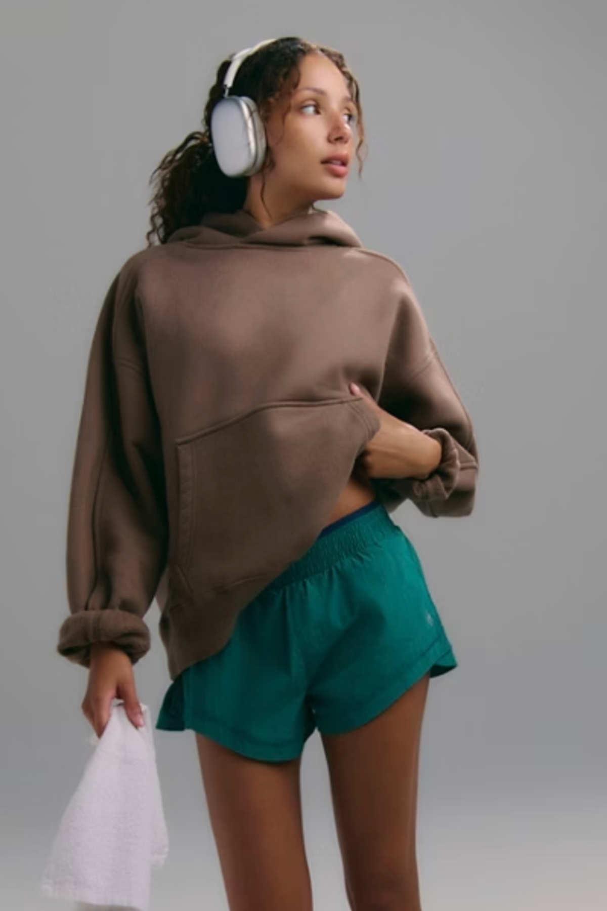 A woman wearing a brown cropped hoodie and teal athletic shorts, with white over-ear headphones and a white towel, standing against a solid gray studio background.