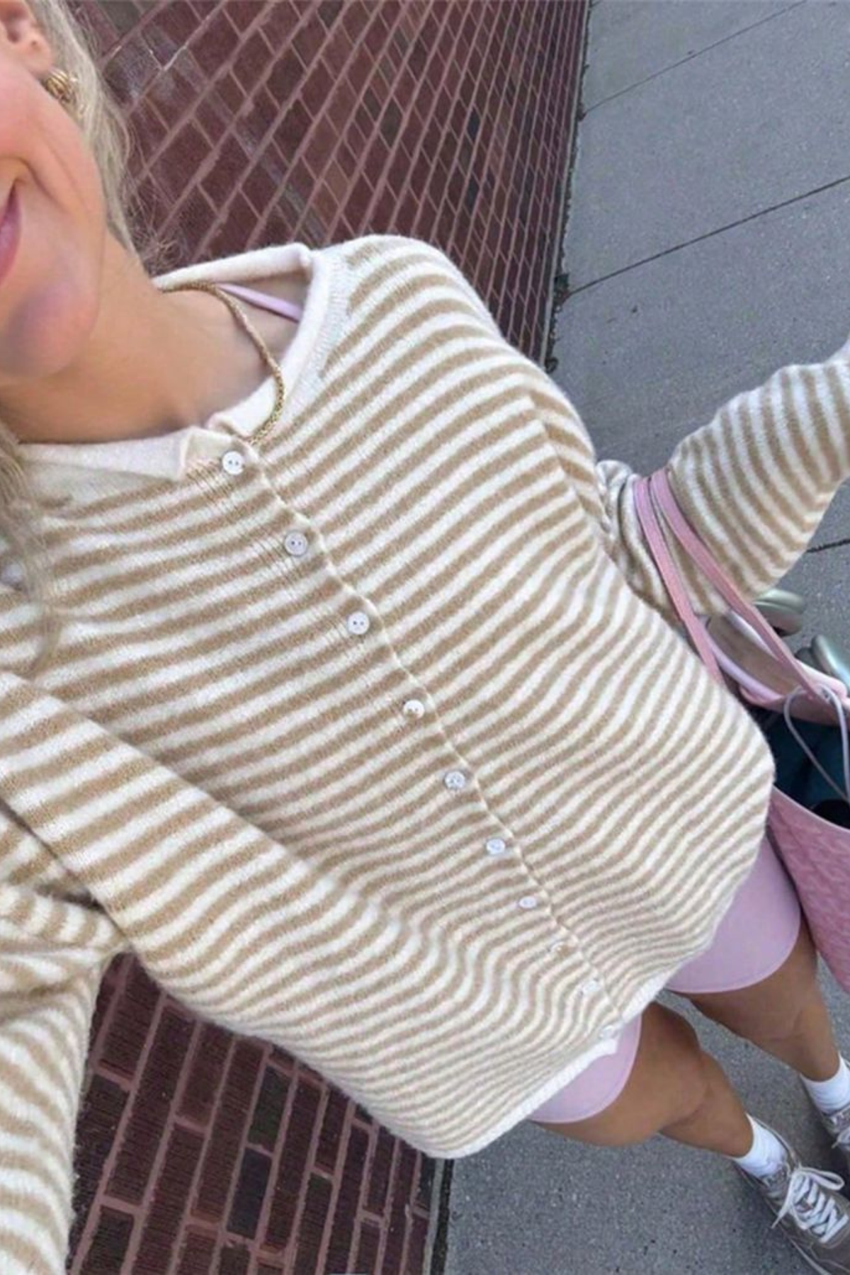 A person wearing a beige and white striped knit cardigan with white buttons, paired with light pink shorts and white sneakers, photographed in a selfie-style close-up on an outdoor sidewalk.