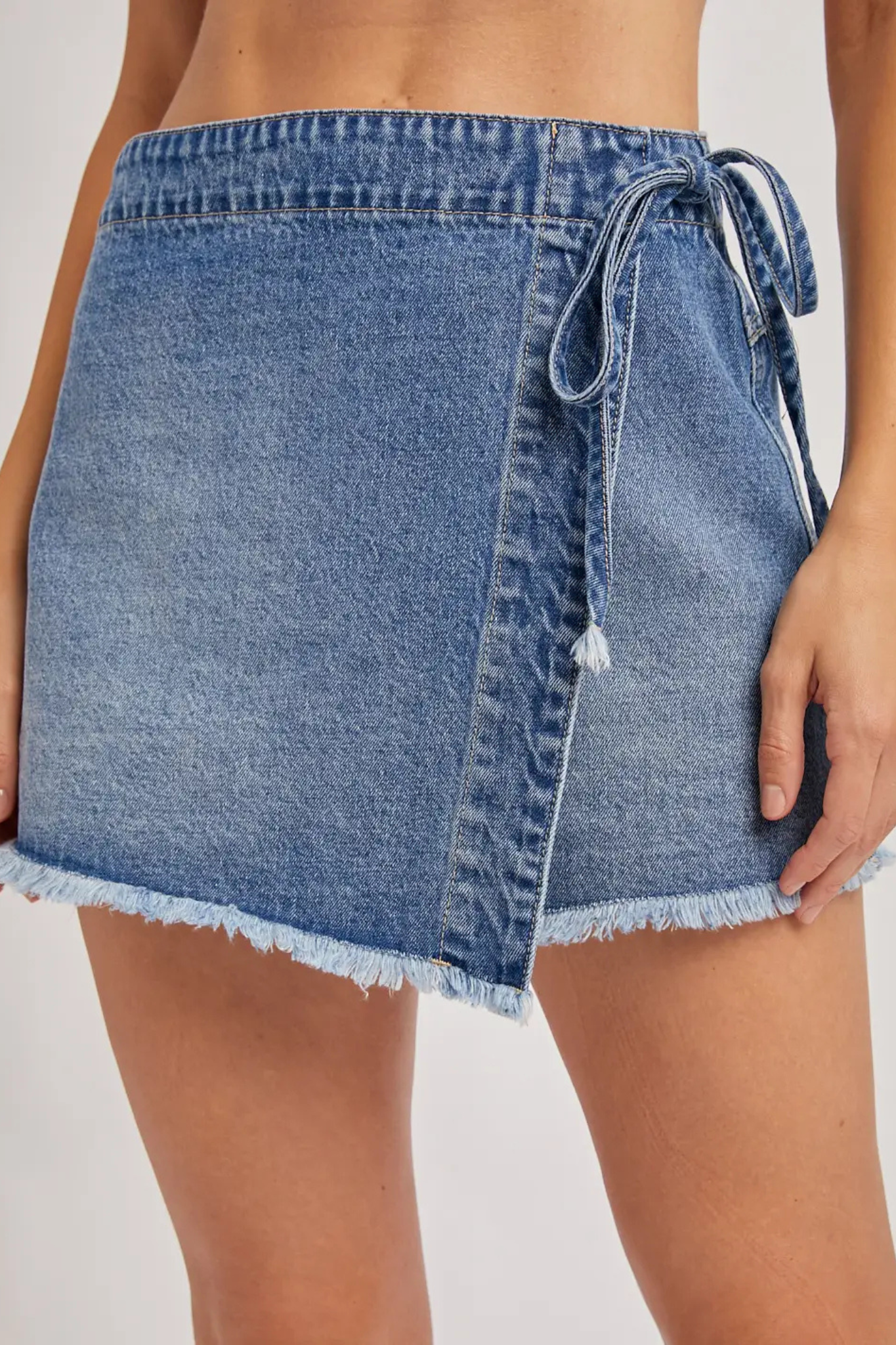 A close-up of a person wearing a light blue denim wrap mini skirt with a side tie and frayed hem, shown against a plain light background.