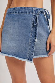 A close-up of a person wearing a light blue denim wrap mini skirt with a side tie and frayed hem, shown against a plain light background.