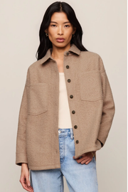 A woman modeling a light brown button-up shirt jacket over a cream top and light blue jeans, set against a plain beige studio background.