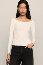 A woman modeling a fitted, off-the-shoulder long-sleeve top in off-white, paired with black jeans, against a plain light gray studio background.