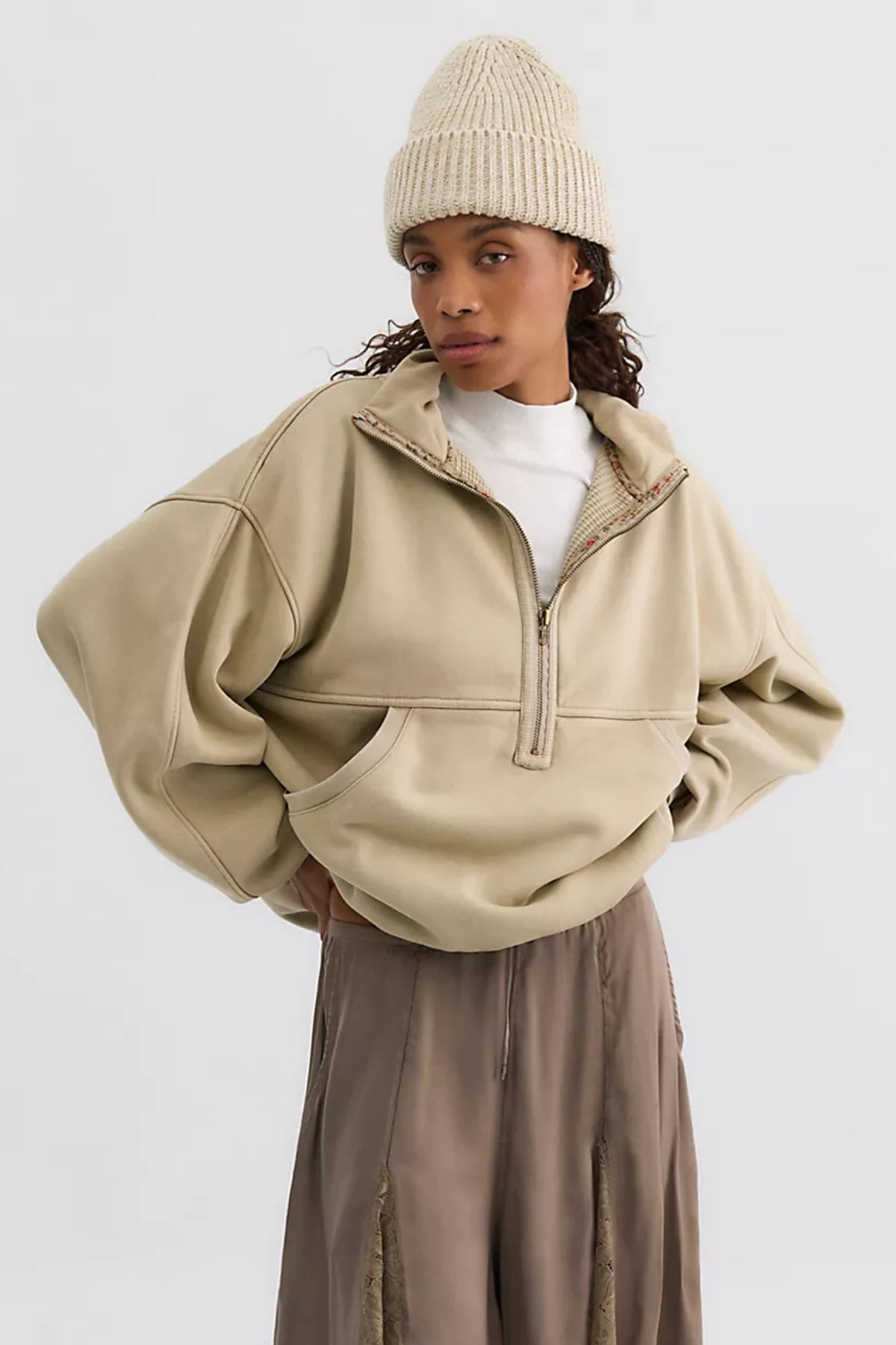 A model wearing a beige oversized quarter-zip pullover jacket with a mock neck, styled with a cream ribbed knit beanie, a white t-shirt, and brown wide-leg trousers, set against a plain light gray studio background.