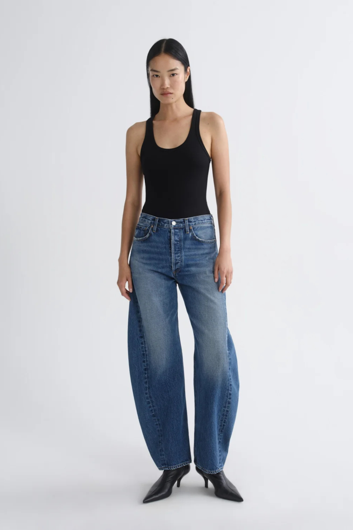 A model stands against a light gray studio background wearing a black ribbed tank top, wide-leg blue denim jeans, and black pointed-toe heels.