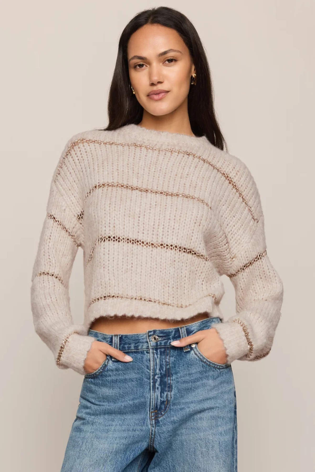 A woman modeling a cropped, fuzzy knit sweater with horizontal brown stripes, paired with blue jeans, against a solid light beige background.
