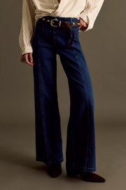 A person wearing dark blue wide-leg denim jeans with a brown leather belt, paired with a cream striped shirt and dark brown pointed-toe shoes, photographed against a plain taupe studio background.