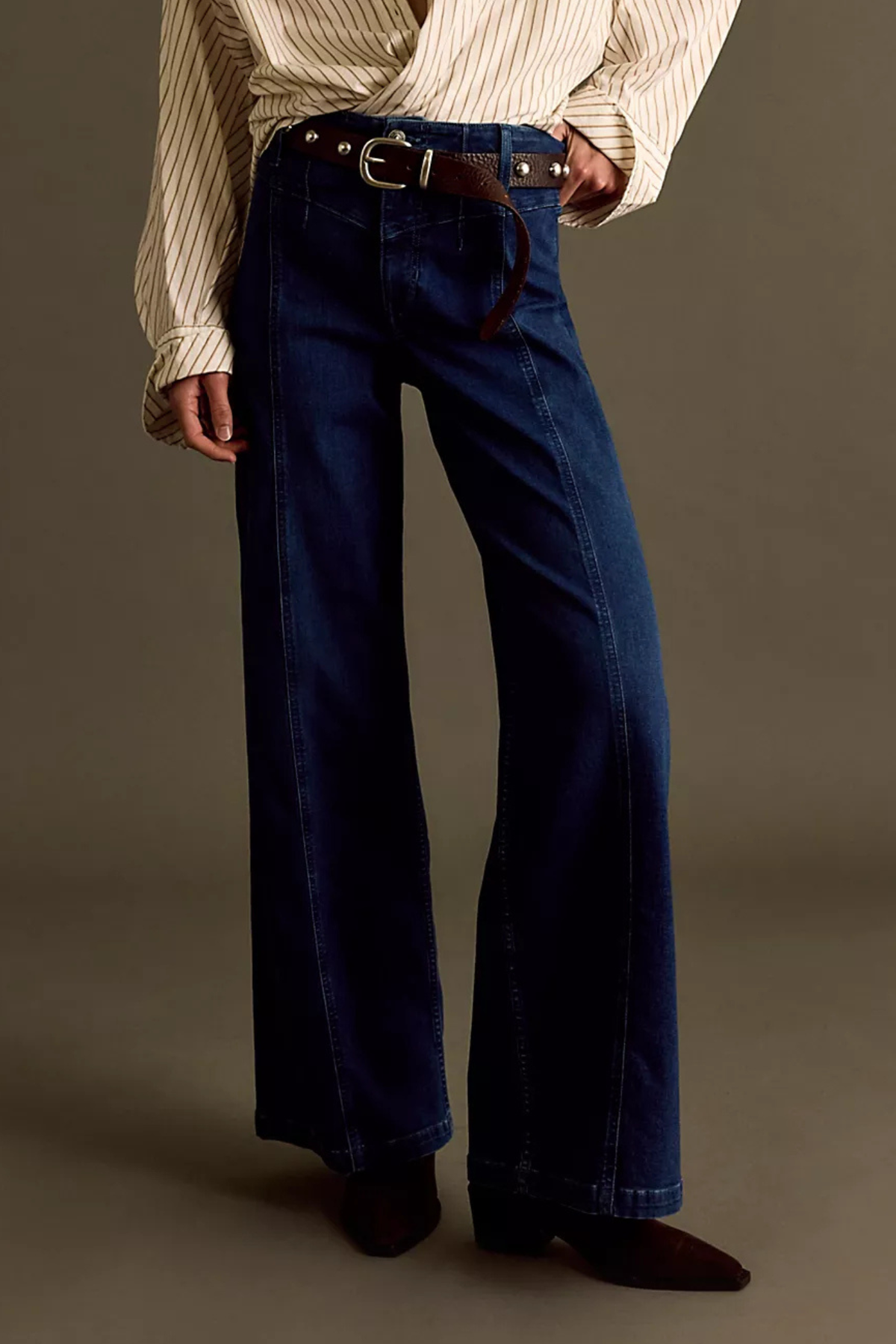 A person wearing dark blue wide-leg denim jeans with a brown leather belt, paired with a cream striped shirt and dark brown pointed-toe shoes, photographed against a plain taupe studio background.