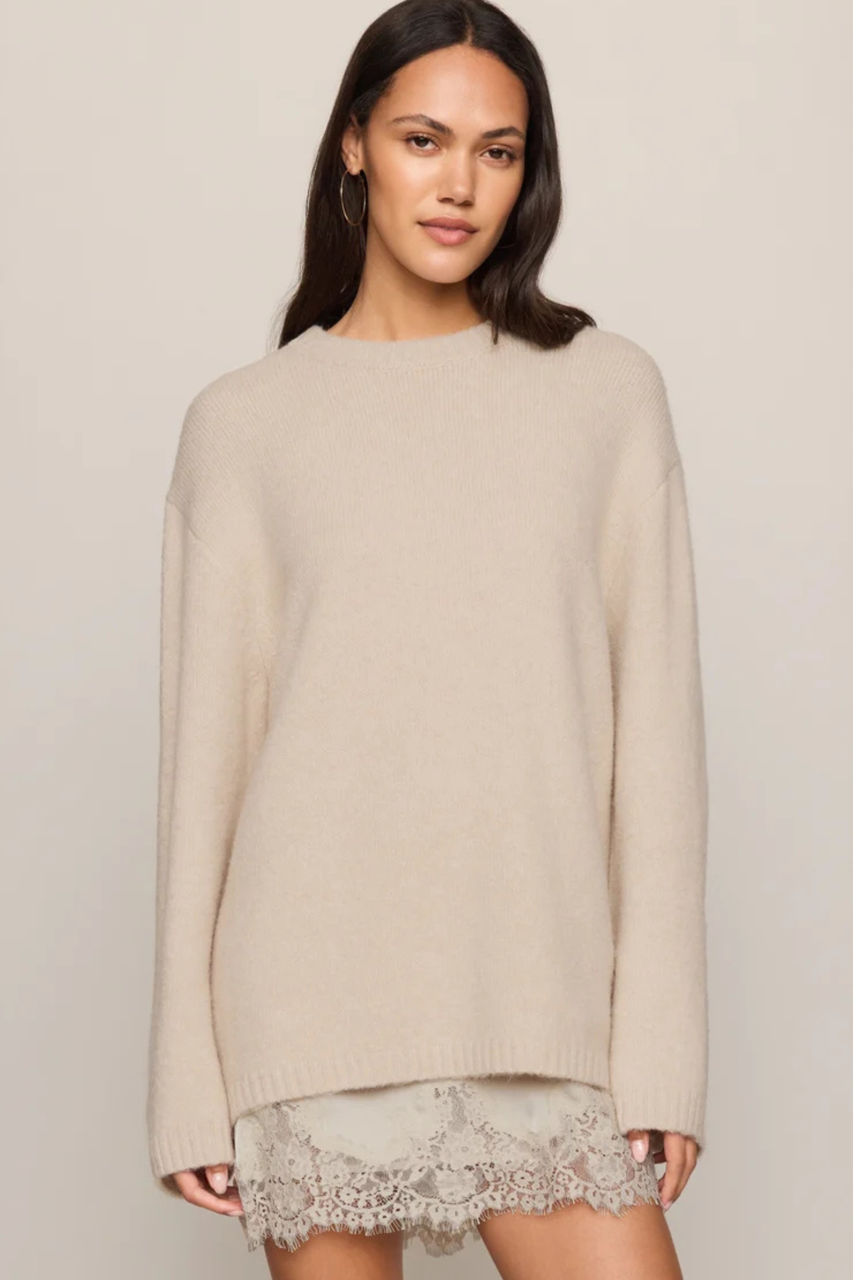 A woman wearing a cream-colored knit sweater with a lace hem, photographed against a plain light beige studio background.
