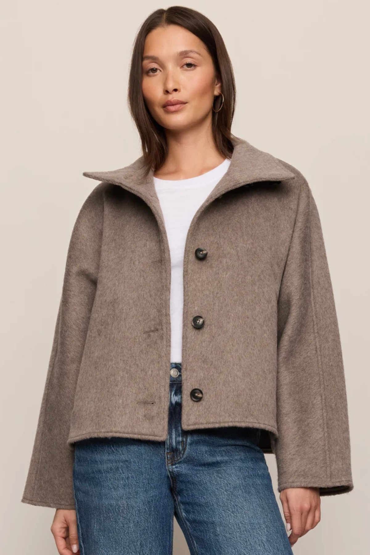 A woman modeling a cropped taupe-brown wool coat with a wide collar and dark buttons, worn over a white top and blue jeans, against a plain beige studio background.