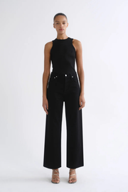 A model stands against a plain white studio background wearing a black sleeveless mock-neck top and matching black wide-leg trousers, paired with beige strappy heels.