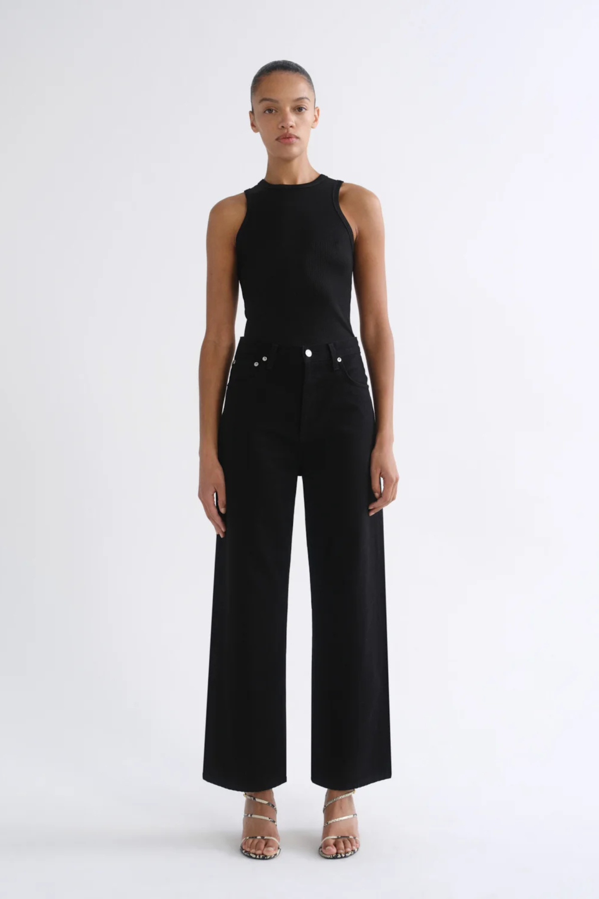 A model stands against a plain white studio background wearing a black sleeveless mock-neck top and matching black wide-leg trousers, paired with beige strappy heels.