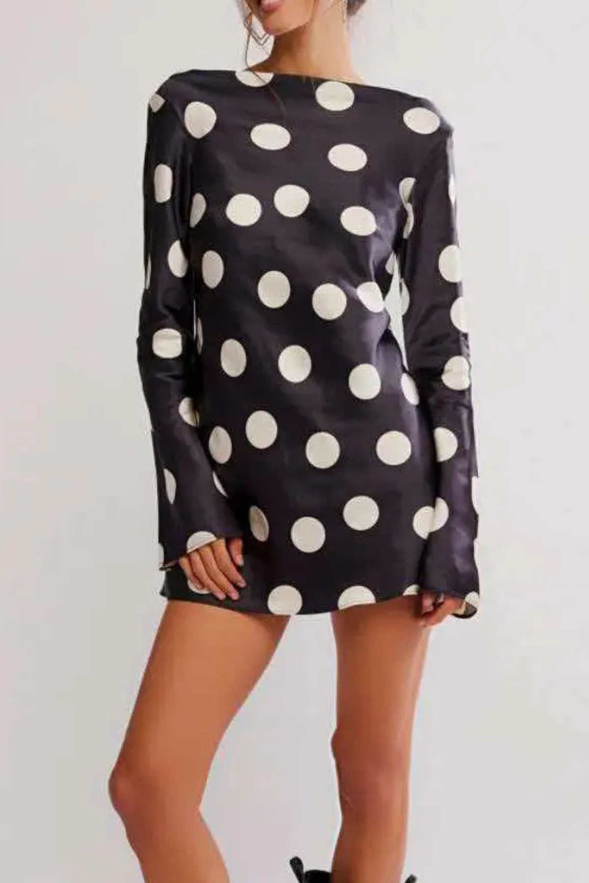 A woman wearing a black satin long-sleeve mini dress with large white polka dots, shown from the neck down against a plain light background.