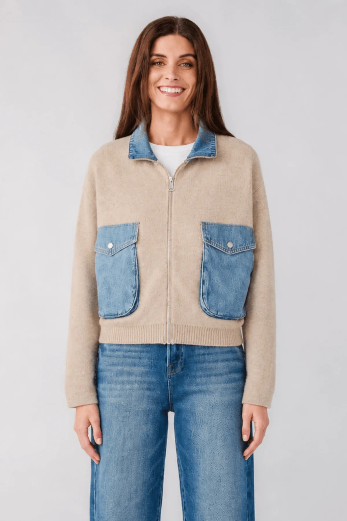 A woman modeling a cropped beige zip-up sweater with blue denim pockets and collar, paired with blue jeans, against a light gray studio background.