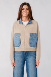 A woman modeling a cropped beige zip-up sweater with blue denim pockets and collar, paired with blue jeans, against a light gray studio background.