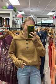 A woman takes a mirror selfie in a clothing boutique wearing a mustard yellow ribbed knit sweater with a buttoned collar and light blue jeans.