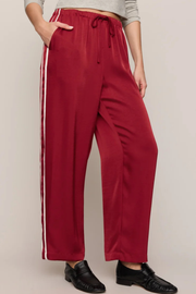 A person modeling wide-leg drawstring pants in deep red with two white side stripes, paired with a light gray cropped top and black loafers, against a plain light beige studio background.