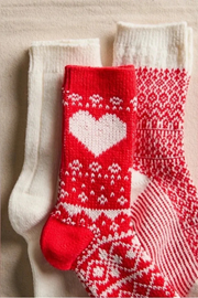 Three knitted socks arranged on a light beige fabric background: one white sock, one red-and-white heart-patterned sock, and one red-and-white Nordic patterned sock.