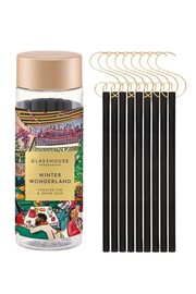 A clear glass jar of Glasshouse Fragrances Winter Wonderland frosted fir and snow gum flavored sticks, with a metallic gold lid and a label featuring a colorful winter illustration. To the right are ten black sticks with gold metal loops at the top, arranged on a plain white background.