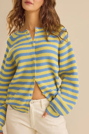 A woman wearing a yellow and light blue horizontally striped ribbed knit cardigan with a buttoned collar and cream pants, photographed against a plain beige studio background.