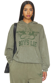 A woman modeling an olive green hooded sweatshirt and matching sweatpants set. The hoodie features a dark green graphic of a winged figure with the text 'BOYS LIE' across the chest.