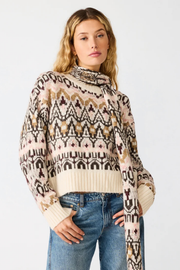 A woman wearing a cream, brown, and black patterned knit sweater with a cowl neck and blue jeans, standing against a light gray studio background.