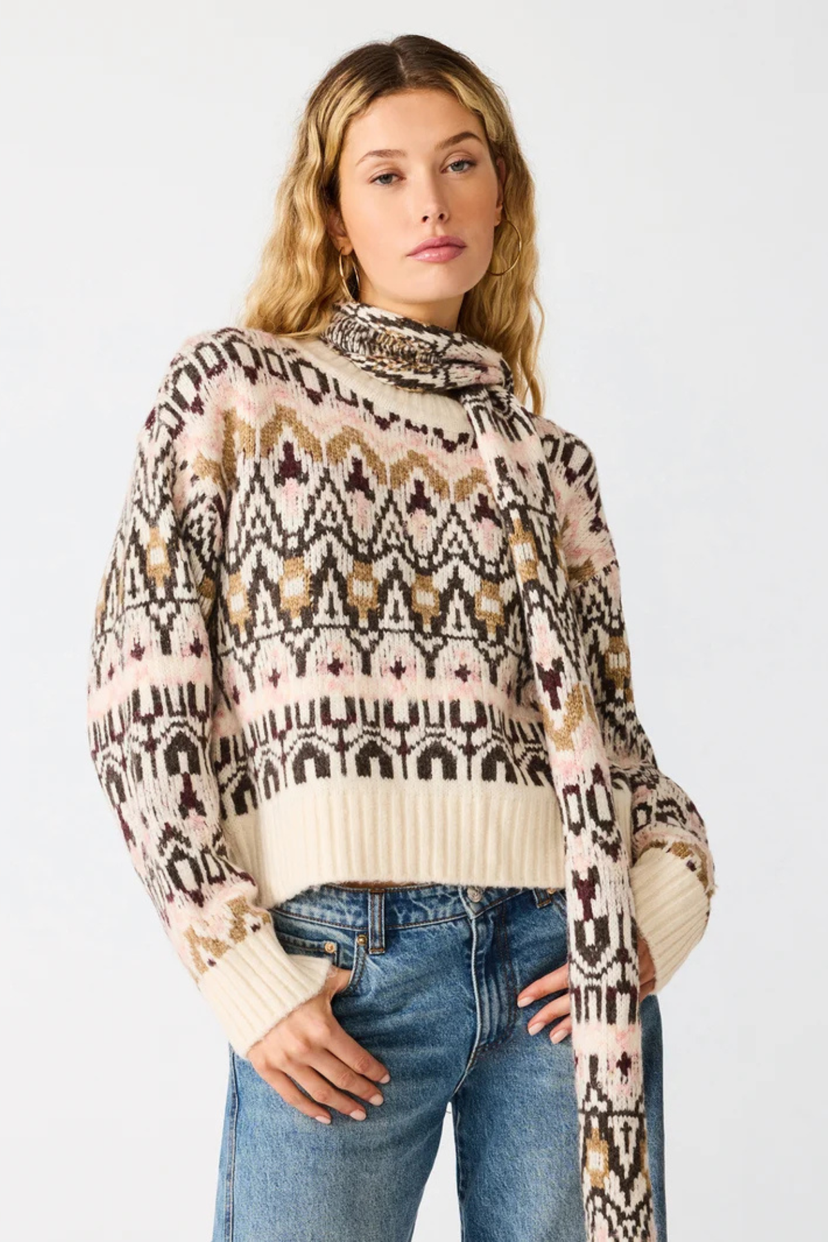 A woman wearing a cream, brown, and black patterned knit sweater with a cowl neck and blue jeans, standing against a light gray studio background.