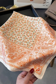 A hand with red nail polish holds a square scarf featuring an orange paisley border and a dense small floral print in orange, green, and red on an off-white background. The scarf is displayed over a dark tray with a beige handbag in the background.