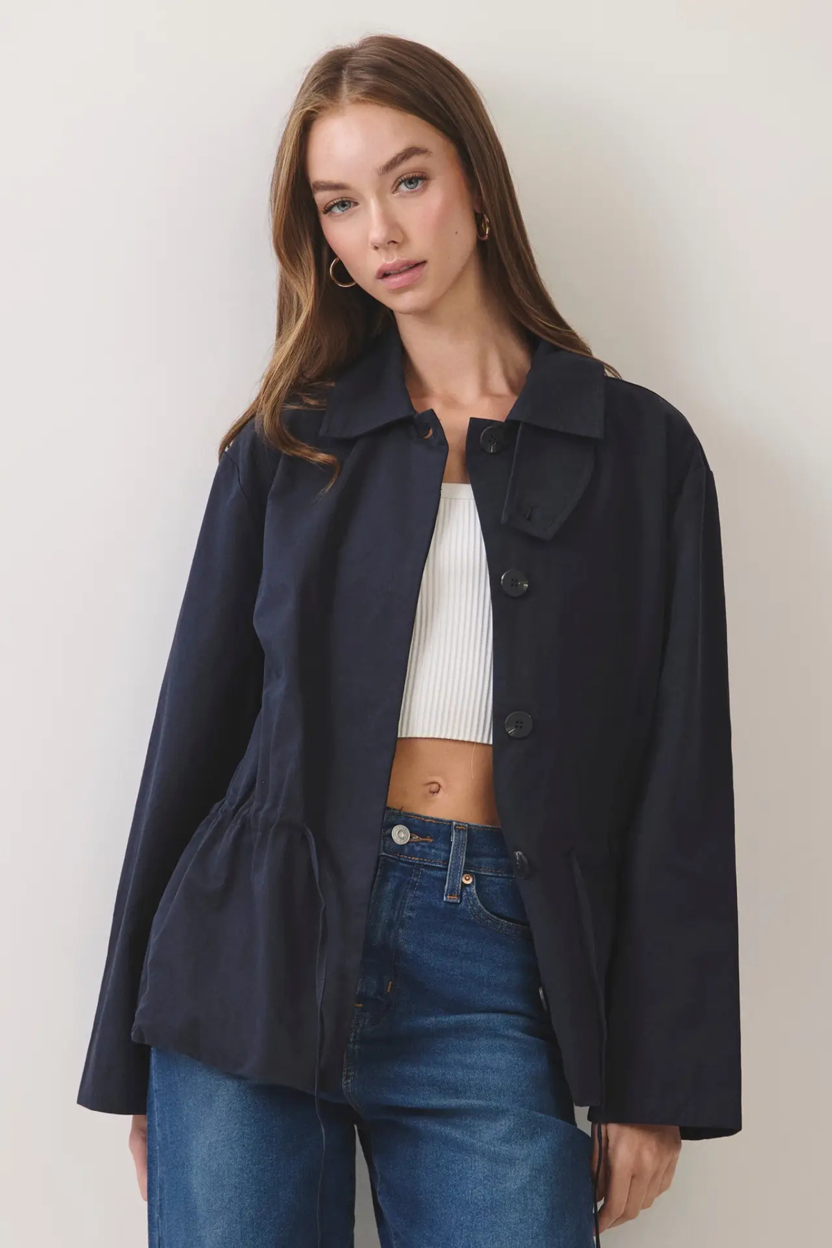 A woman modeling a dark navy blue longline jacket with a collar, front buttons, and a peplum waist, worn over a white ribbed crop top and blue jeans against a plain light background.