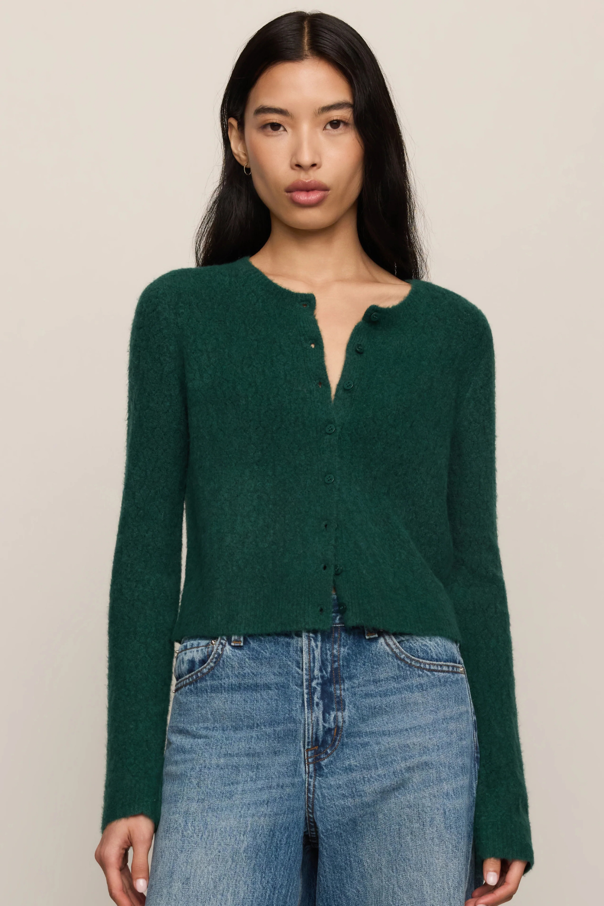 A woman modeling a dark green fuzzy knit cardigan with a split neckline and button details, paired with blue jeans, against a solid light beige background.