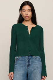 A woman modeling a dark green fuzzy knit cardigan with a split neckline and button details, paired with blue jeans, against a solid light beige background.