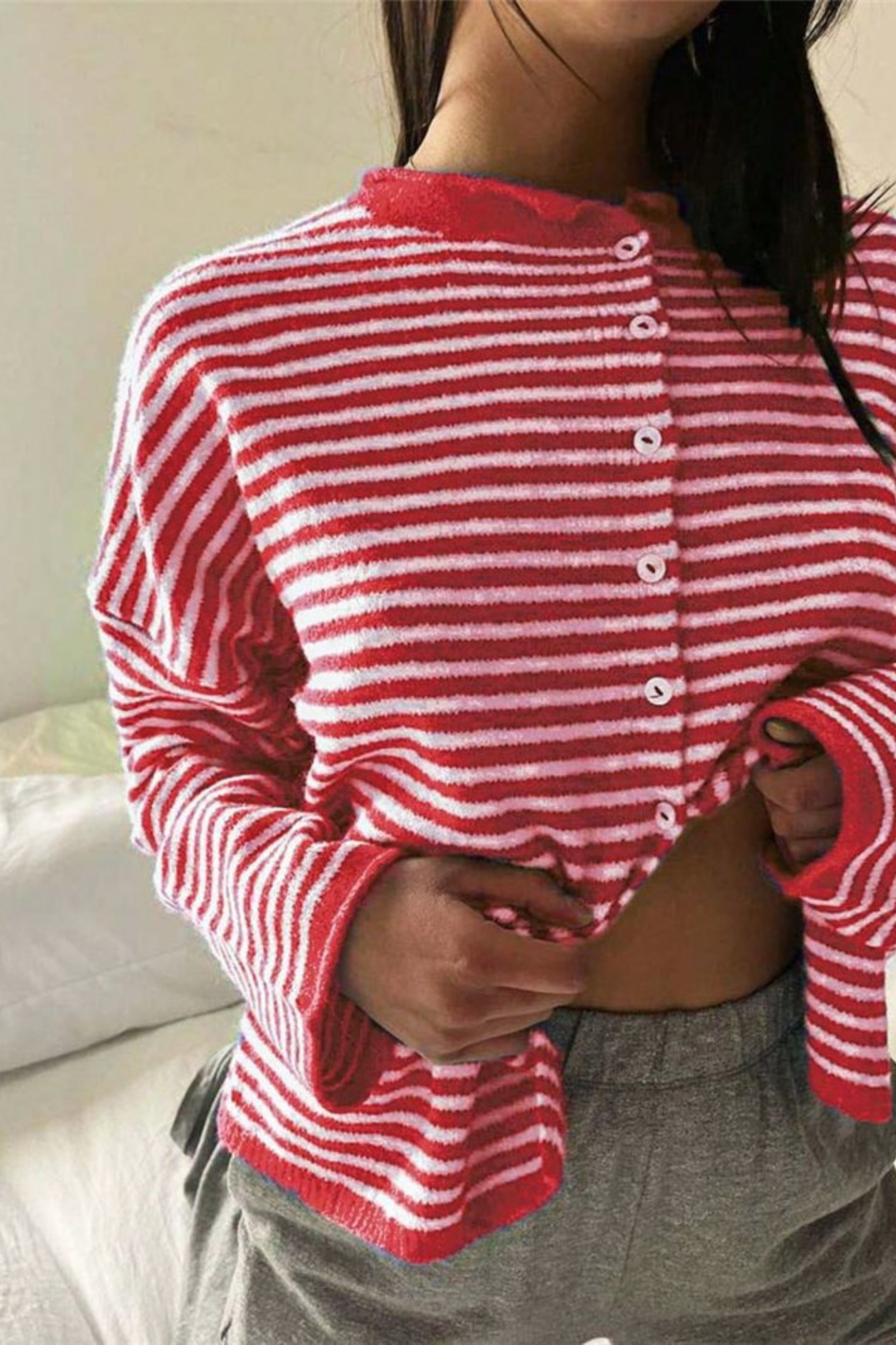 A person wearing a red and white striped long-sleeve cropped cardigan with white buttons, paired with gray sweatpants, in a bedroom setting.