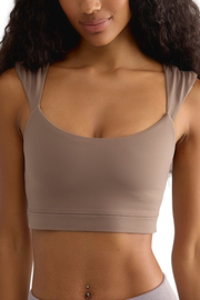 A close-up of a woman wearing a scoop-neck athletic sports bra in a muted taupe color, showing the fit and straps against a plain white background.