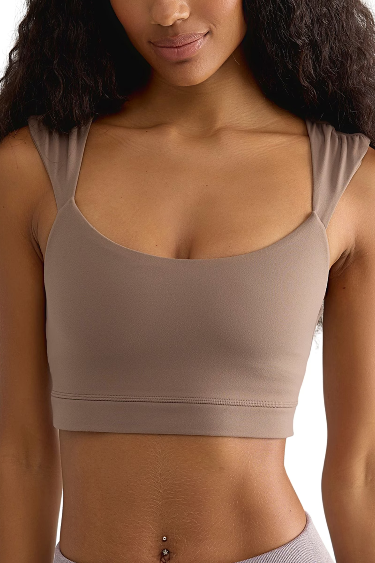 A close-up of a woman wearing a scoop-neck athletic sports bra in a muted taupe color, showing the fit and straps against a plain white background.