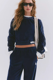 A woman modeling a navy blue velour tracksuit with white piping details, consisting of a cropped long-sleeve top and matching sweatpants, styled with a white shoulder bag and sunglasses against a light gray background.