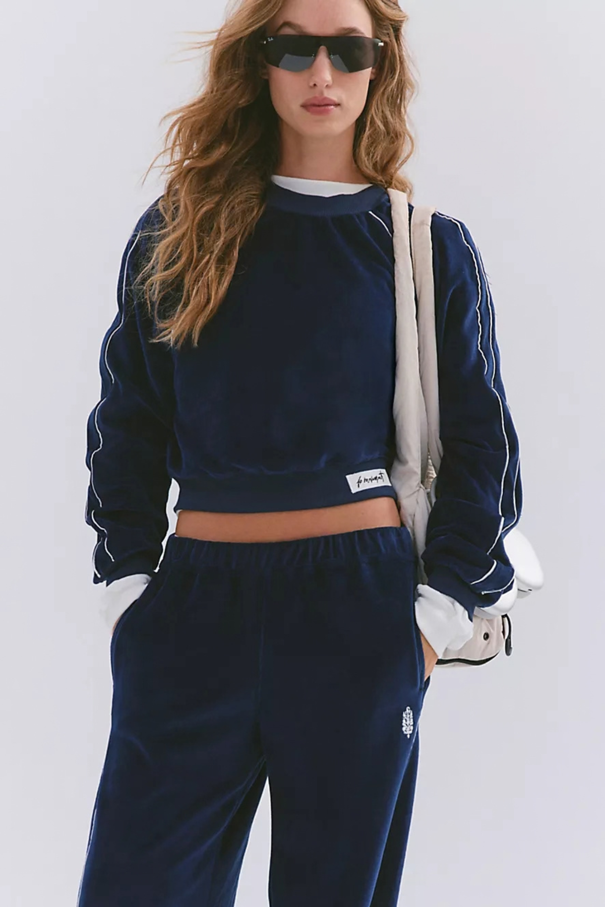 A woman modeling a navy blue velour tracksuit with white piping details, consisting of a cropped long-sleeve top and matching sweatpants, styled with a white shoulder bag and sunglasses against a light gray background.