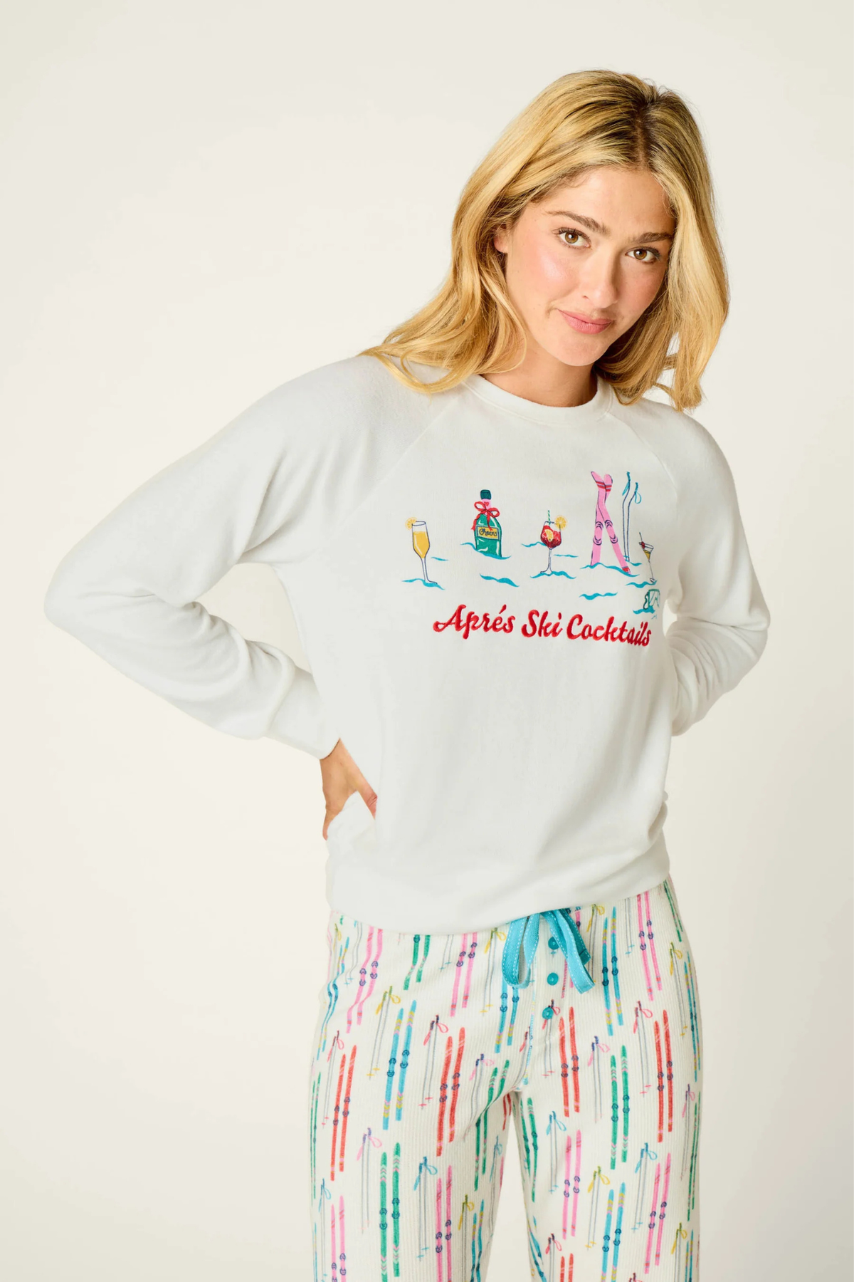 A woman modeling a white long-sleeve crewneck sweatshirt with a colorful graphic of ski-themed cocktails and the text 'Apré Ski Cocktails', paired with multicolored tie-dye pajama pants against a plain off-white background.