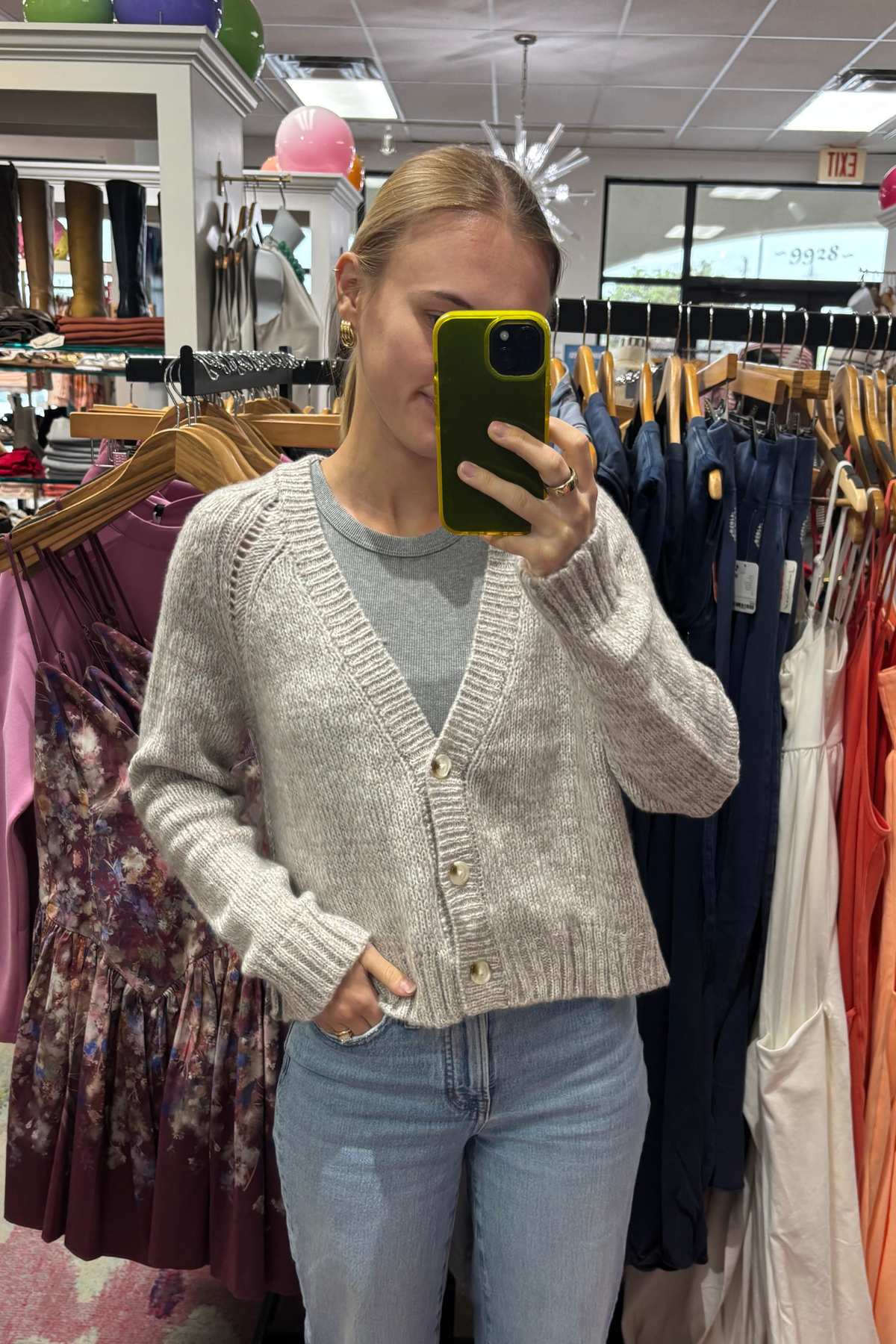 A woman takes a mirror selfie in a clothing store while wearing a light beige knit button-up cardigan over a gray t-shirt and light blue jeans.