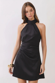 A woman modeling a black satin halter mini dress with a high mock neck and gathered skirt details, set against a plain light gray studio background.