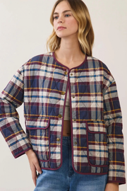 A woman wearing a quilted plaid jacket with navy, burgundy, yellow, and white stripes, paired with blue jeans, against a plain off-white background.