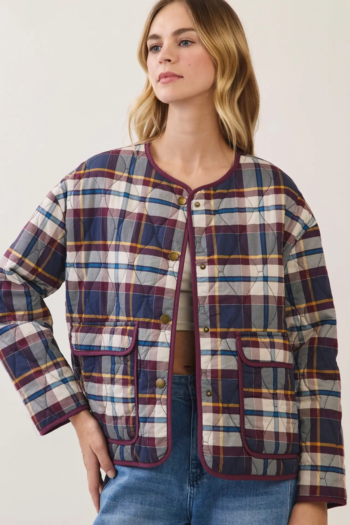 A woman wearing a quilted plaid jacket with navy, burgundy, yellow, and white stripes, paired with blue jeans, against a plain off-white background.